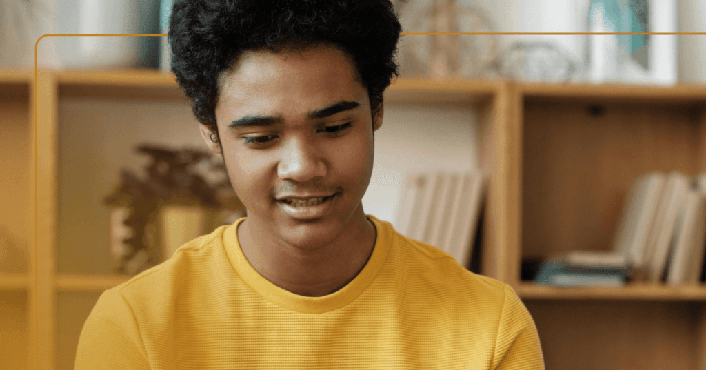 Young man sits staring down at the floor in front of him. His teeth are visible as if he is about to speak. He is wearing a yellow long sleeve shirt. The background is a tan bookshelf.