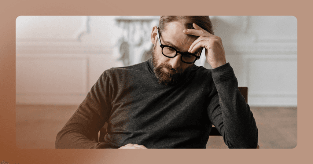 Man sits at a table looking down. He is holding his head with his left hand and his facial expression is serious. He is wearing black glasses and a turtleneck. The background is a white wall. There is a brown border around the image.