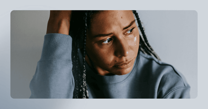 Woman sits staring to the left with a tear coming down her cheek. She is grabbing her head with her right hand and her facial expression is sad. She is wearing a blue crewneck and the background is a gray wall.