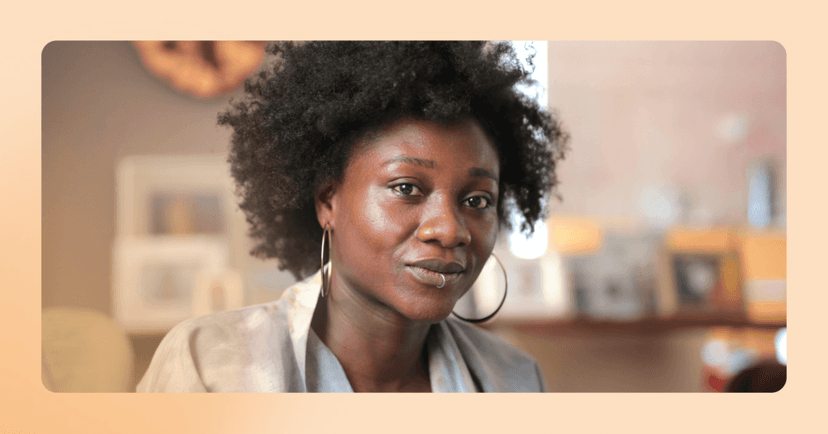 Woman looks directly into camera with a serious, unsure look. She has dark brown curly hair, is wearing a blazer and is in front of a blurred bookshelf in the background.