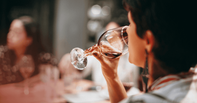 A woman drinks red wine from wine glass. She is in focus in the foreground and two other people sitting at the table are blurred in the background.