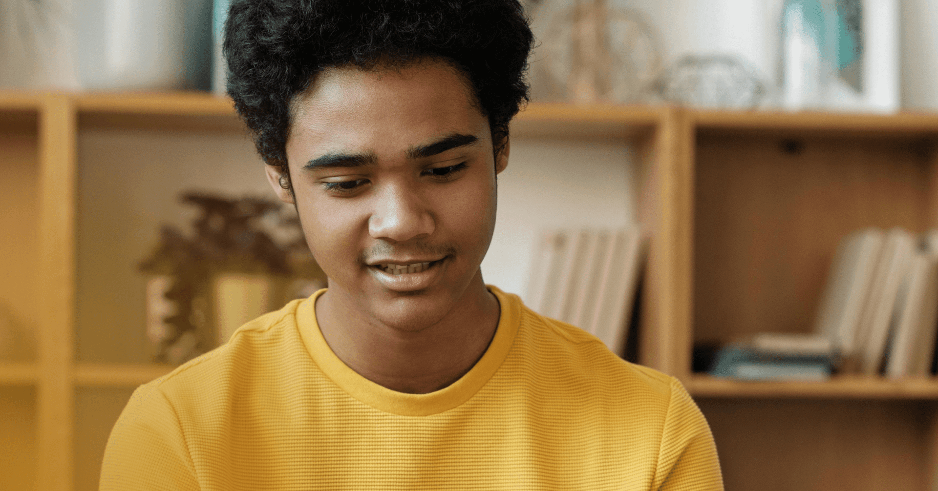 A male teenager is looking straight down off camera. There is a bookshelf visible in the background. He is wearing wearing a yellow shirt.