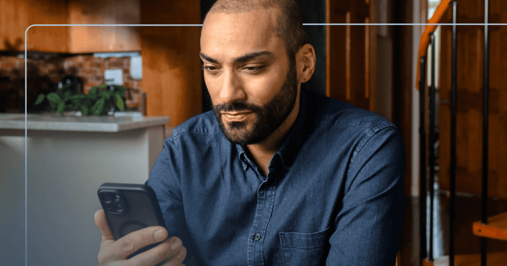 Man sits at a wooden table in his home staring down at his phone in his right hand. His facial expression is neutral. He is wearing a blue denim button up shirt. The background is a kitchen to the left and a staircase on the right.