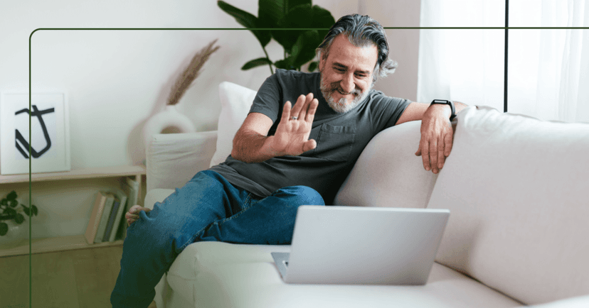Older man sits on a white couch with an open laptop in front of him. He is waving his right hand and smiling looking down at the laptop screen. He is wearing a dark green T-shirt and jeans. The background is a bookshelf against a white wall and a window to the right.