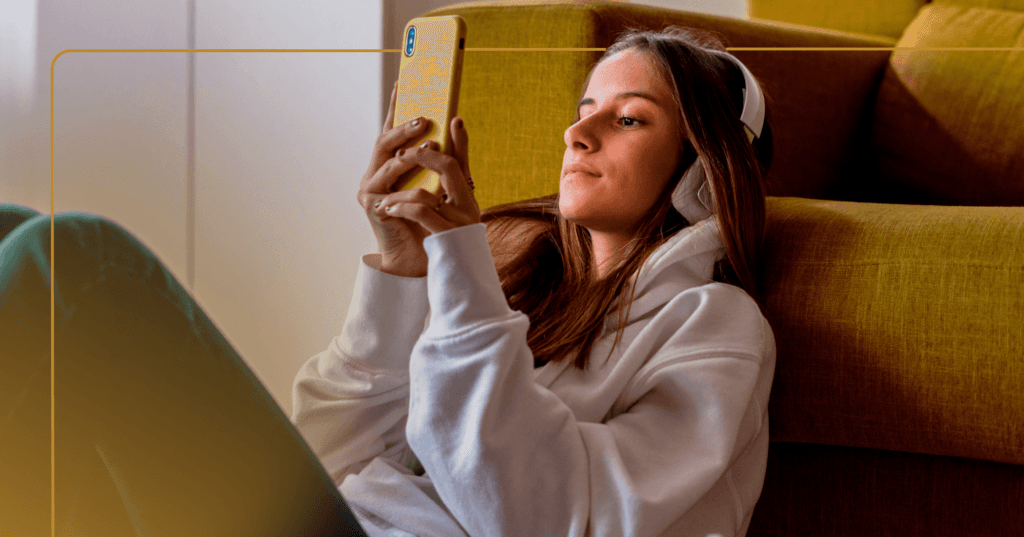 Young girl sits on the floor leaning against a yellow couch. She is looking at the yellow phone she holds in front of her in both hands. She is wearing white over the ear headphones and a white hoodie. There is a yellow border around the image.