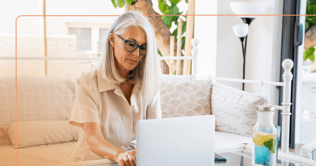 Older woman sits on a white couch with an open white laptop in front of her on a glass table. She is typing and looking down at the laptop screen. She is wearing a beige shirt and blue framed glasses. The background is a sliding glass door and a lamp.