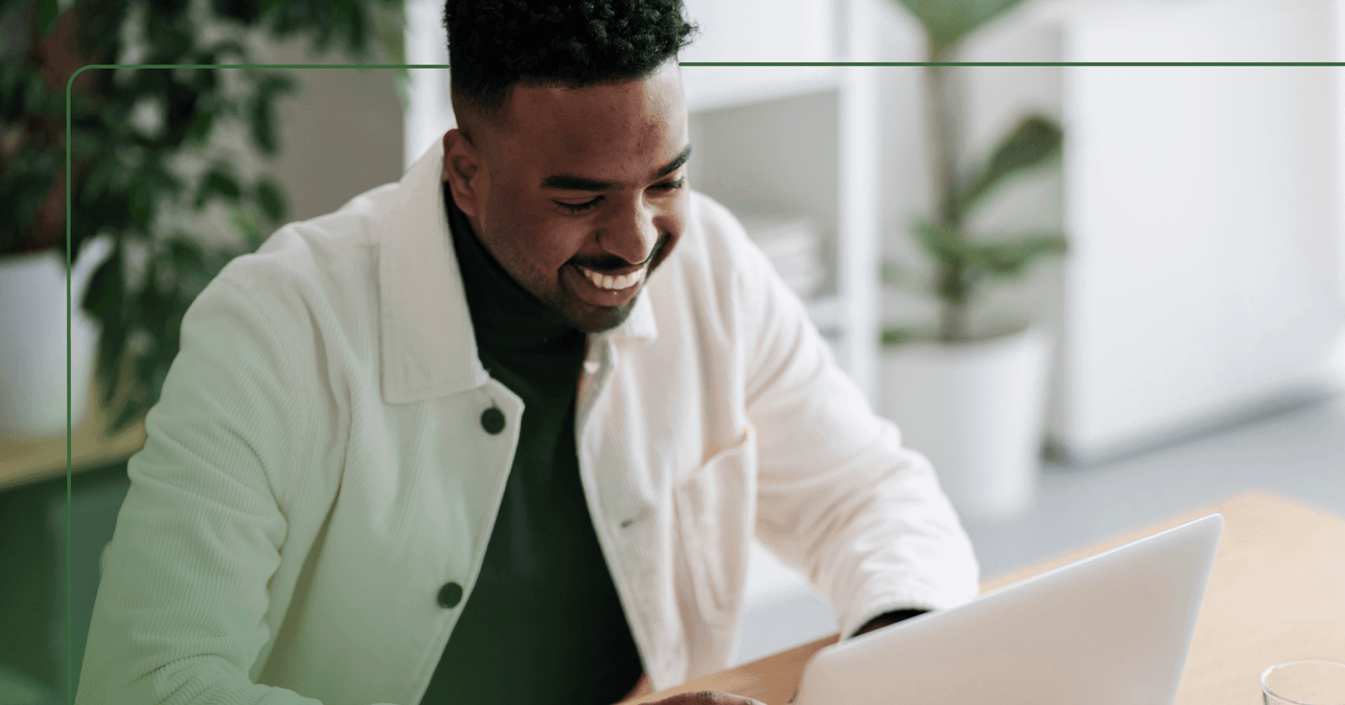 Man in a white button down shirt looks down at a laptop while smiling. He is sitting at a wooden table with plants of our focus in the background.