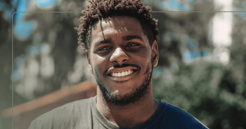 Man stands outside staring straight at the camera. He is smiling with all his teeth and the sun is shining. He is wearing a black T-shirt. The background is trees that are blurry.