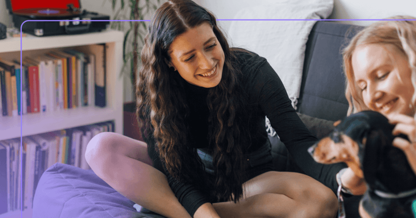 Two young women sit on a couch petting a dog in the right corner of the screen. One woman has brown hair and is sitting with her legs crossed. The other woman has blonde hair and her body is not visible behind the dog. Both of their facial expressions are happy and smiling at the dog. The background is a record player on a full bookshelf.