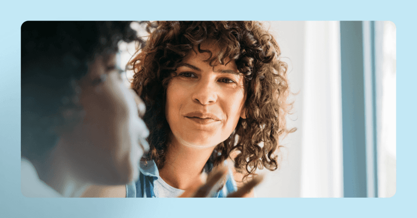 Woman is staring at another blurred person who is talking. Her facial expression is interested. She has curly hair. The background is blue and white.