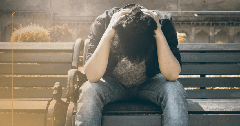 Young man sits outside on a park bench. He has both hands behind his head and his head is facing down. His facial expression is not visible. He is wearing a black button-up shirt over a gray shirt with blue jeans. The background is trees.