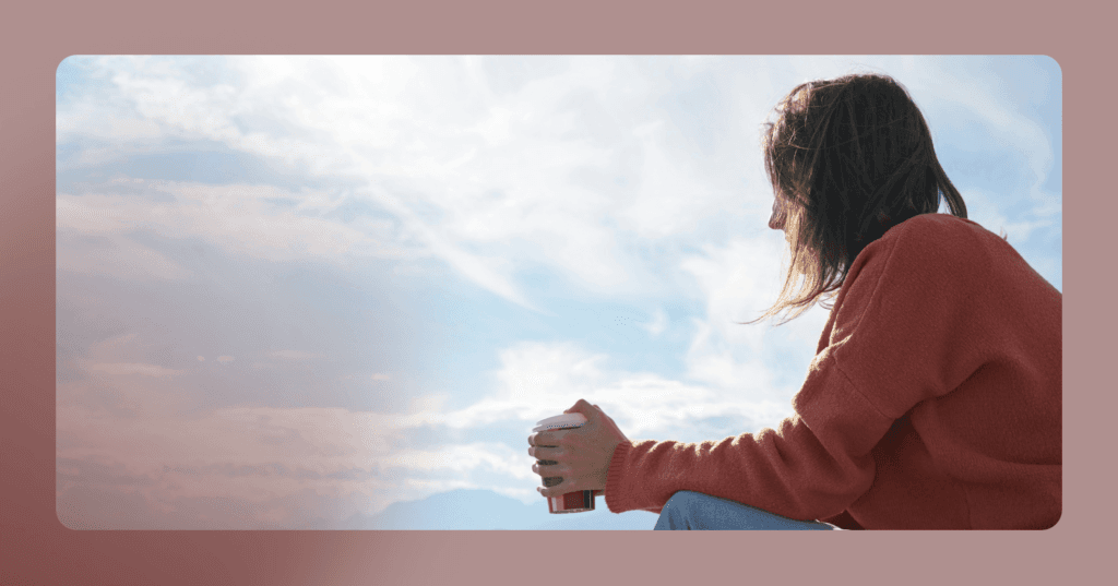 A woman in an orange sweatshirts holds a coffee to-go cup and looks of the frame to the left. A blue sky with clouds is visible in the background. The image is framed in a light red color.