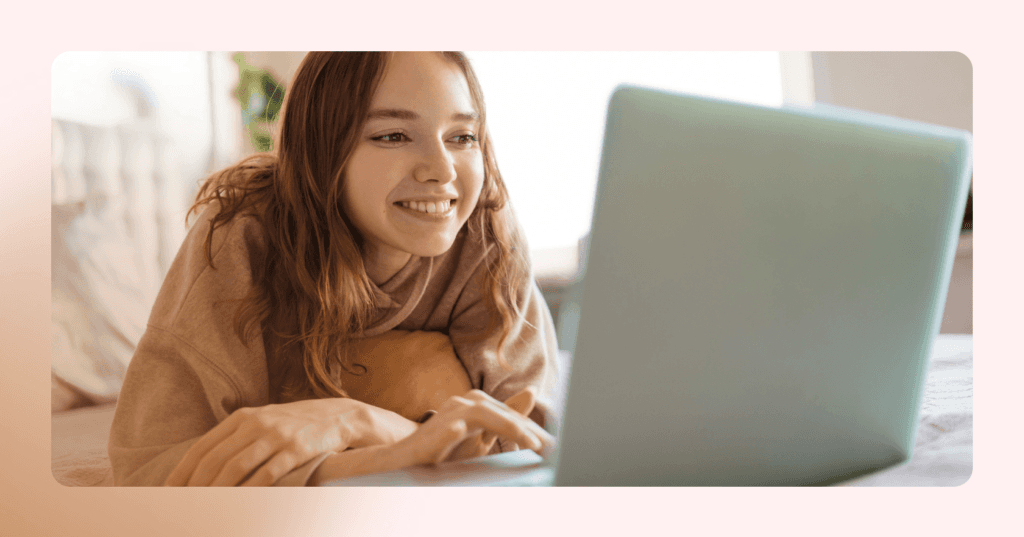 Young girl is lying on her stomach on her bed scrolling on her laptop. She is smiling and staring at the laptop screen. She is wearing a brown pullover sweater. The background is a white bedroom.