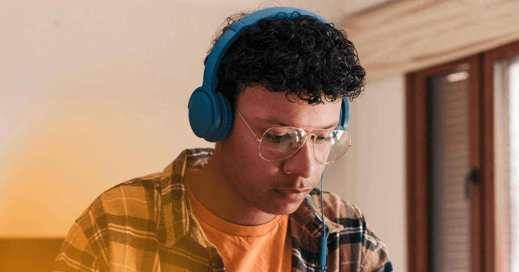Man sits wearing blue over the ear headphones looking down at something offscreen. He is wearing an orange T-shirt underneath a flannel shirt and glasses. The background is a white wall.