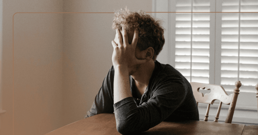Man sits at a wooden table inside his home. His right hand is covering his face. He is wearing a dark gray long sleeve shirt. The background is white shutters and a white wall.