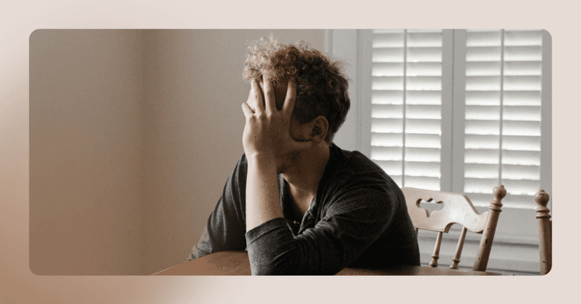 Man sits at a wooden table inside his home. His right hand is covering his face. He is wearing a dark gray long sleeve shirt. The background is white shutters and a white wall.