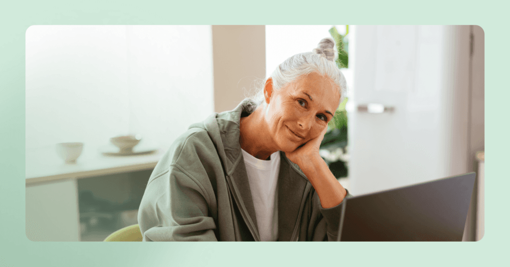Woman sits in front of laptop at table, head resting in one hand. She looks directly at the camera. The image is framed in green.