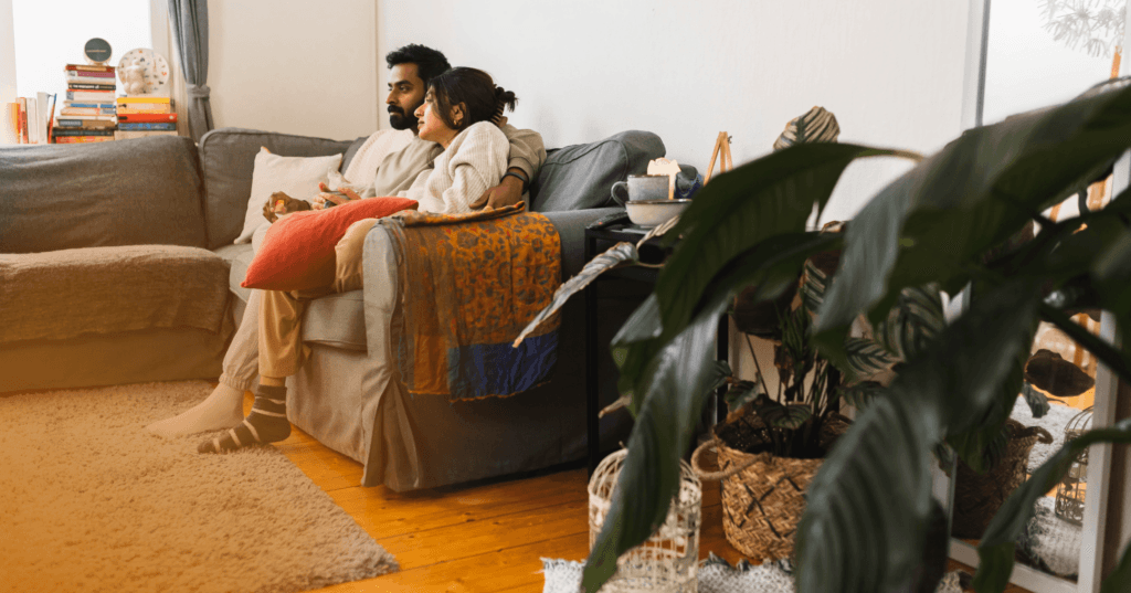 Couple sits together on a dark gray couch. His arm is around her as she holds a mug and they are looking forward perhaps at a TV. He is wearing a light green crewneck sweater with gray sweats and she is wearing a light gray sweater with tan pants. They are inside a home.