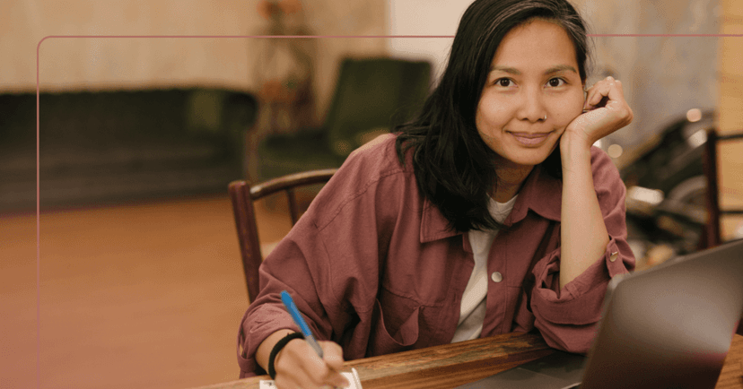 Woman sits at a wooden table with an open laptop in front of her staring straight into the camera. She is smiling and leaning on her left hand. She is wearing a white T-shirt underneath a red undershirt as she writes with a blue pen in her right hand. The background is an empty green chair and couch.