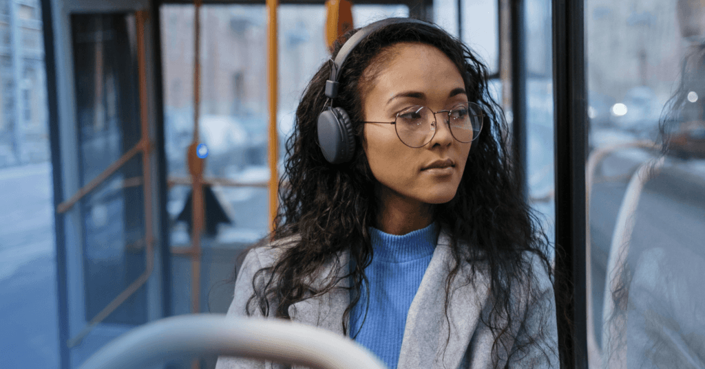 Woman sits on a bus staring out the window on her left. Her facial expression is neutral. She is wearing black over the ear headphones, a blue turtleneck, and gray coat. The background is an open bus door.