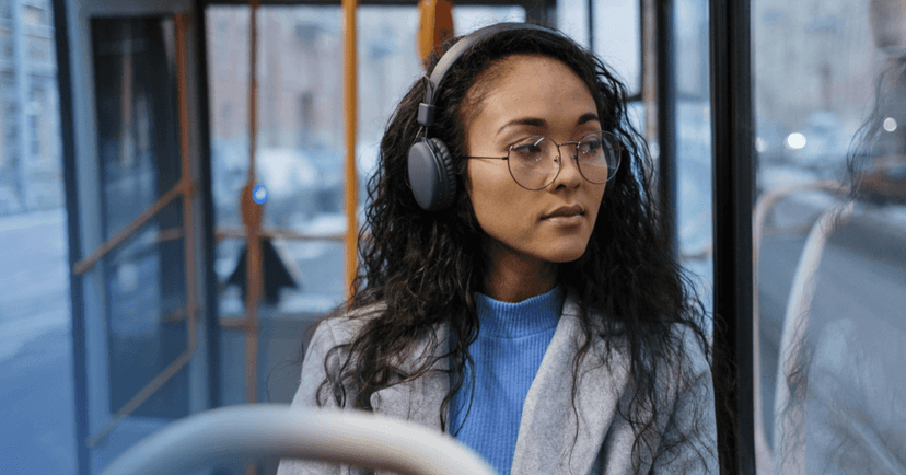 Woman sits on a bus staring out the window on her left. Her facial expression is neutral. She is wearing black over the ear headphones, a blue turtleneck, and gray coat. The background is an open bus door.