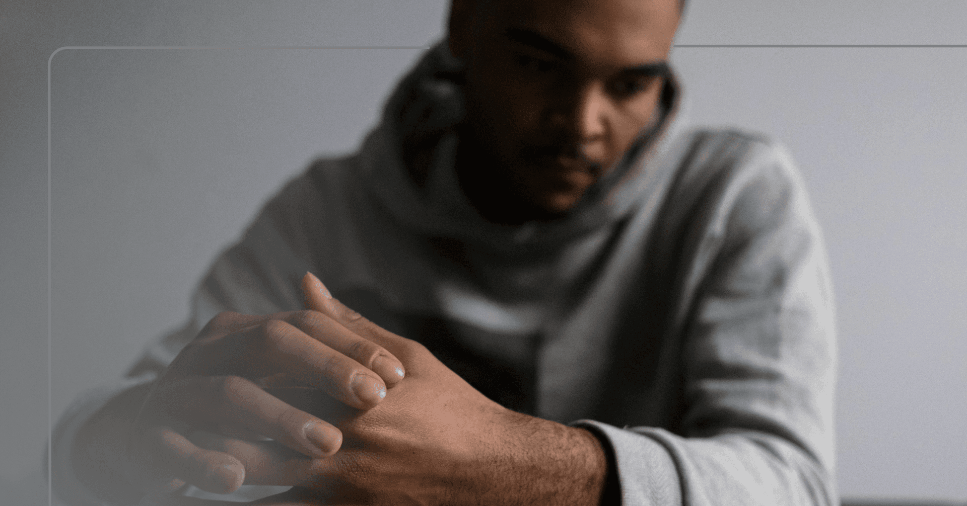 A man in a grey hooded sweatshirt sits with his hands held together. He looks off camera to the left. There is a gray wall in the background.