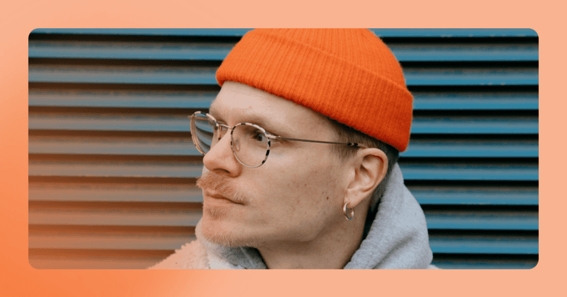 Man with orange beanie and glasses looks off camera to the right. He is standing against a blue metal textured wall. There is an orange frame around the image.