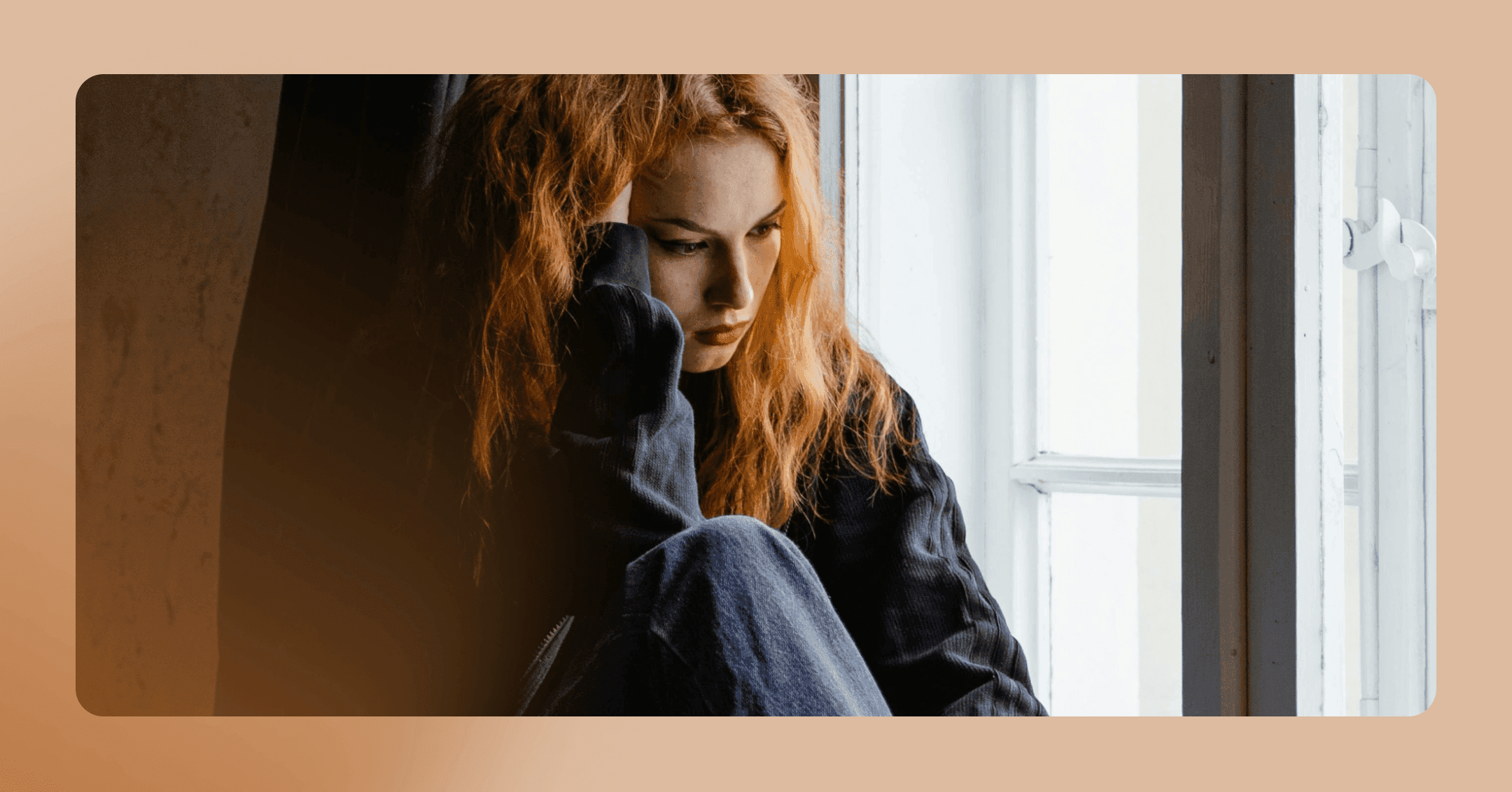 Woman sits next to a window where the light shines on her face. She is looking down and holding her head with her right hand. Her facial expression is concerned. She is wearing a black sweatshirt and jeans. The background is a wall. There is an orange border around the image.