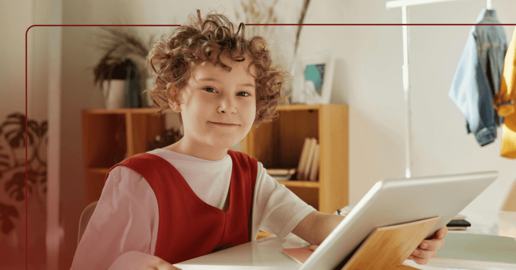 Child sits at a desk in their bedroom with a tablet on a wooden stand in front of them. They are staring at the camera and grinning. They are wearing a red and white shirt. The background is a wooden bookshelf against a white wall next to a jean jacket hanging on a metal rack.