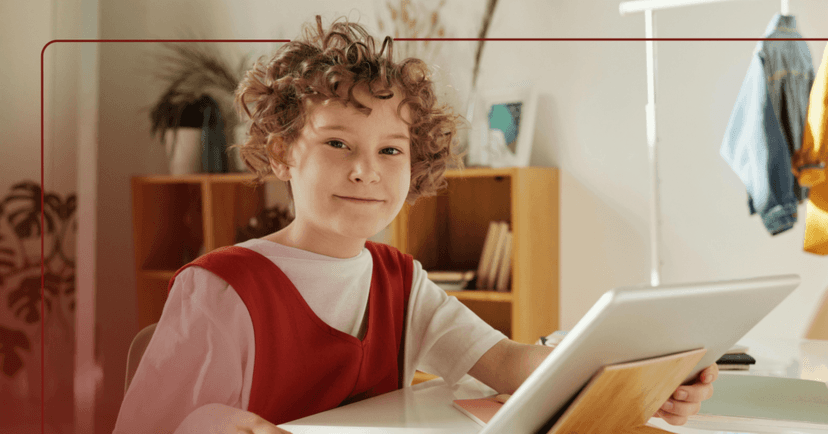 Child sits at a desk in their bedroom with a tablet on a wooden stand in front of them. They are staring at the camera and grinning. They are wearing a red and white shirt. The background is a wooden bookshelf against a white wall next to a jean jacket hanging on a metal rack.
