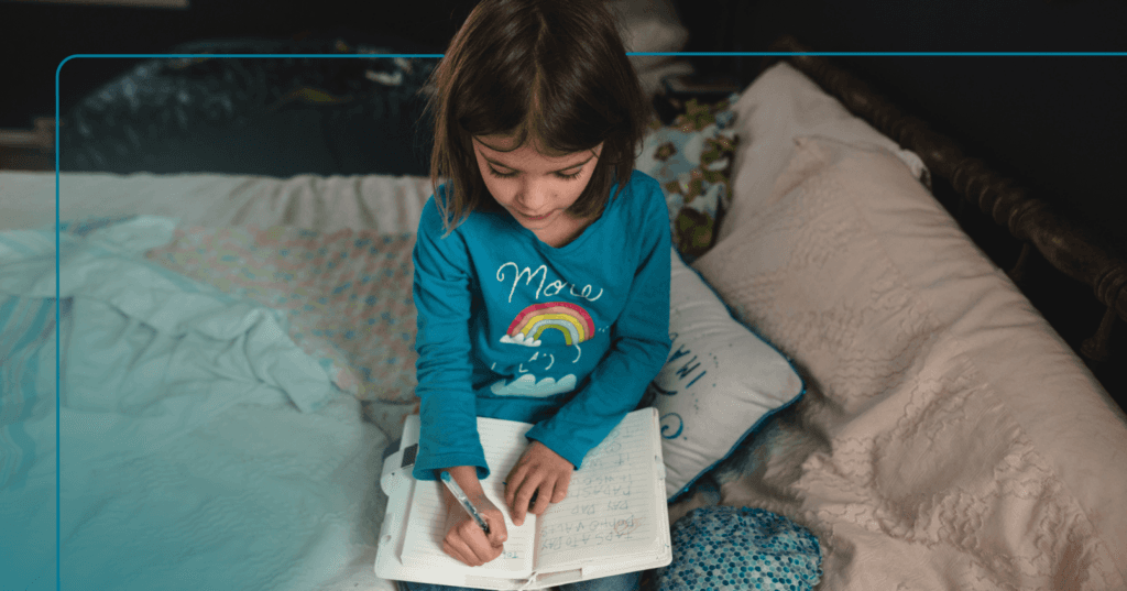Child sits on a bed writing with a pen in a notebook. She is looking down at the paper and is focused. She is wearing a blue long sleeve shirt with a rainbow on it. The background is a bedroom.