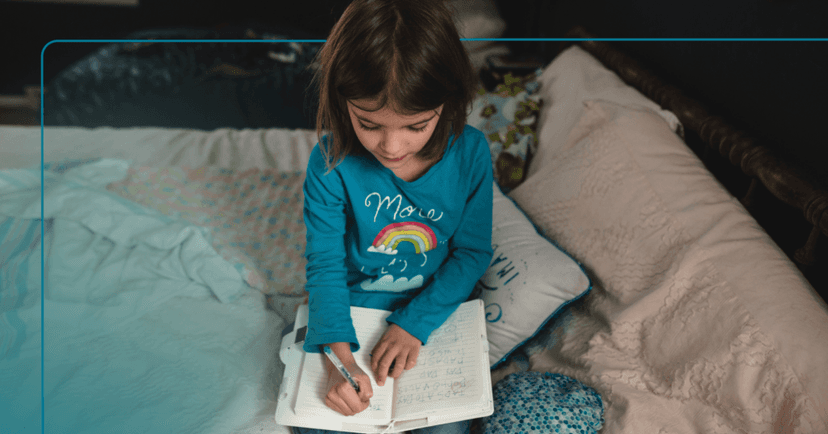 Child sits on a bed writing with a pen in a notebook. She is looking down at the paper and is focused. She is wearing a blue long sleeve shirt with a rainbow on it. The background is a bedroom.