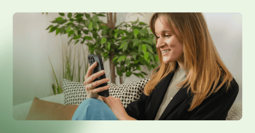 Woman sits on a couch looking down at her phone. She is holding her phone in a black case with her right hand and smiling at the screen. She is wearing a black blazer over a green shirt and jeans. The background is two pillows next to her on the couch and two plants.