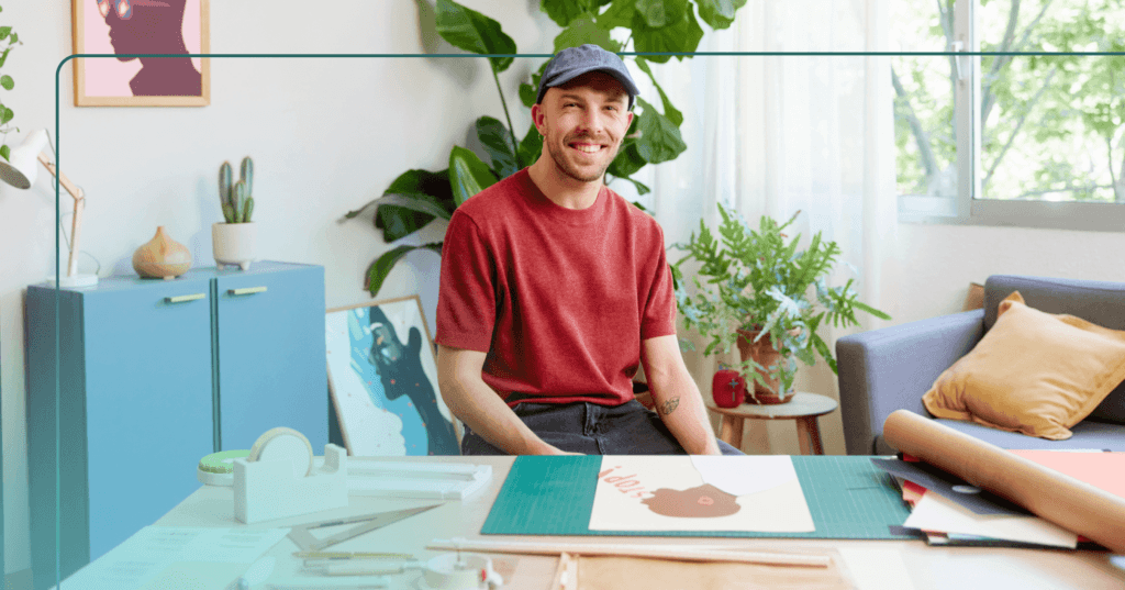Man sits at a wooden table indoors staring straight at the camera. He is smiling and has art supplies on the table in front of him. He is wearing a blue cap, red T-shirt, and black jeans. The background is a blue cabinet on the left, two green plants, and a gray couch in front of a window.