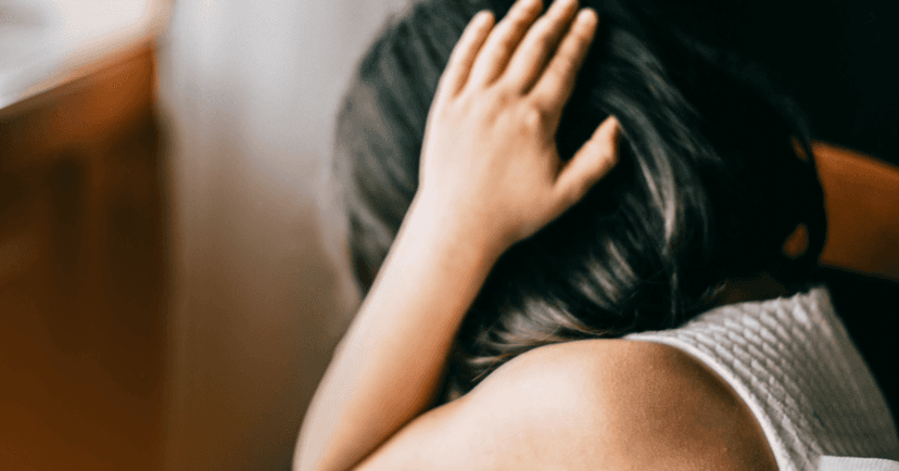 Young child sits with her hands covering her ears. Her facial expression is not visible. She is wearing a white blouse and her head is pointed down. The background is a wooden chair.