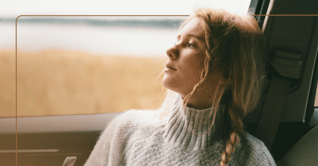 Girl sits in a car staring out the window on her right. Her facial expression is distracted. She is wearing a gray turtleneck and has two braids in her hair. The background is a lake.