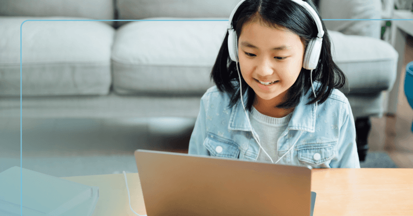 Young girl sits on the floor at a long wooden table. She is looking down at an open laptop in front of her smiling with her teeth. She is wearing a gray sweater underneath a jean jacket with white over the ear headphones. The background is a gray couch.