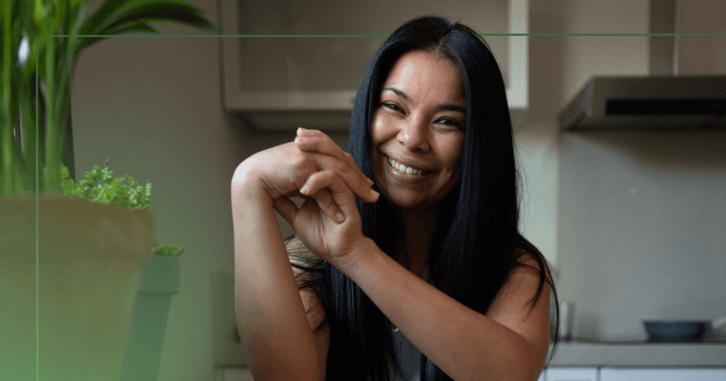 Woman sits at a kitchen table staring straight at the camera. She is clasping her hands together and smiling with her teeth. She has black hair and is sitting beside two plants. The background is a kitchen.