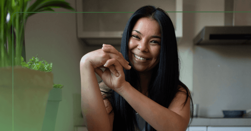 Woman sits at a kitchen table staring straight at the camera. She is clasping her hands together and smiling with her teeth. She has black hair and is sitting beside two plants. The background is a kitchen.