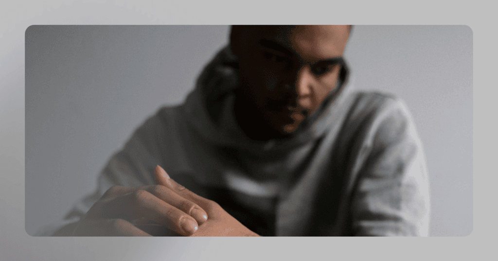 Man sits with his hands together looking down to the left in front of him. His facial expression is serious. He is wearing a gray hoodie against a gray wall in the background. The image is blurry and sharper in focus on the left side.