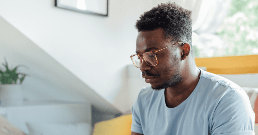 Man sits on a couch staring down in front of him. His facial expression is serious and unfocused. He is wearing glasses and a blue T-shirt. The background is a white wall and an open window.