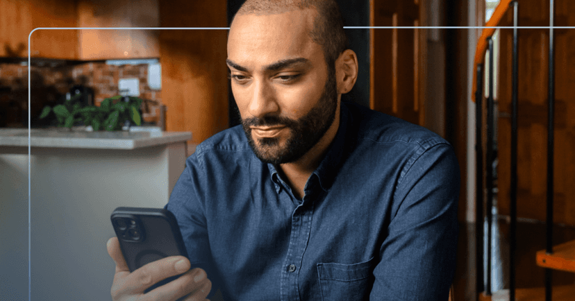 Man sits at a wooden table in his home staring down at his phone in his right hand. His facial expression is neutral. He is wearing a blue denim button up shirt. The background is a kitchen to the left and a staircase on the right.