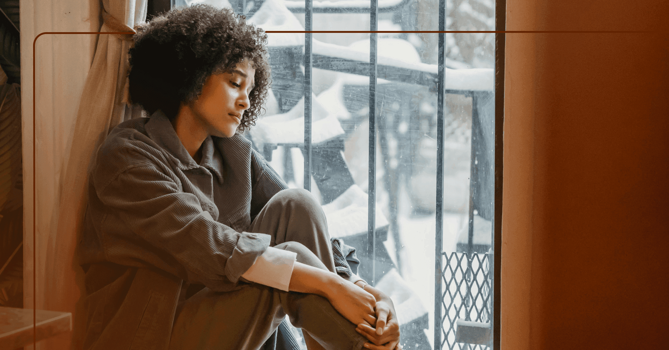 Woman sits on the floor next to a window. She is holding her bent knees and looking down. Her facial expression is sad. She is wearing a gray long sleeve shirt and gray pants. There are black metal bars on the window.