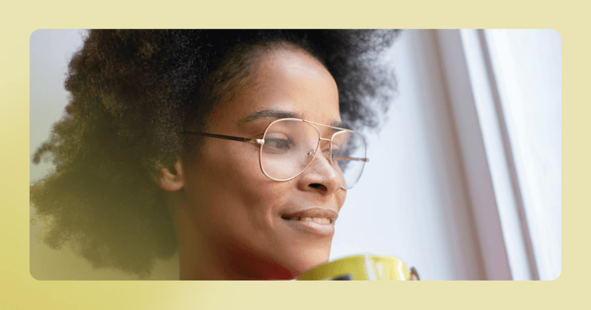 Woman with glasses sits against a wall with a cup of coffee. She is looking off camera. The image is framed in light yellow.