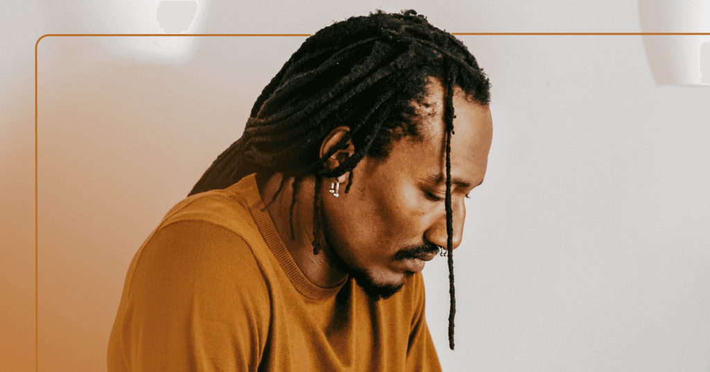 Man with dreadlocks and a brown shirt looking down and to the right. He has a sad face. The image is framed in brown.