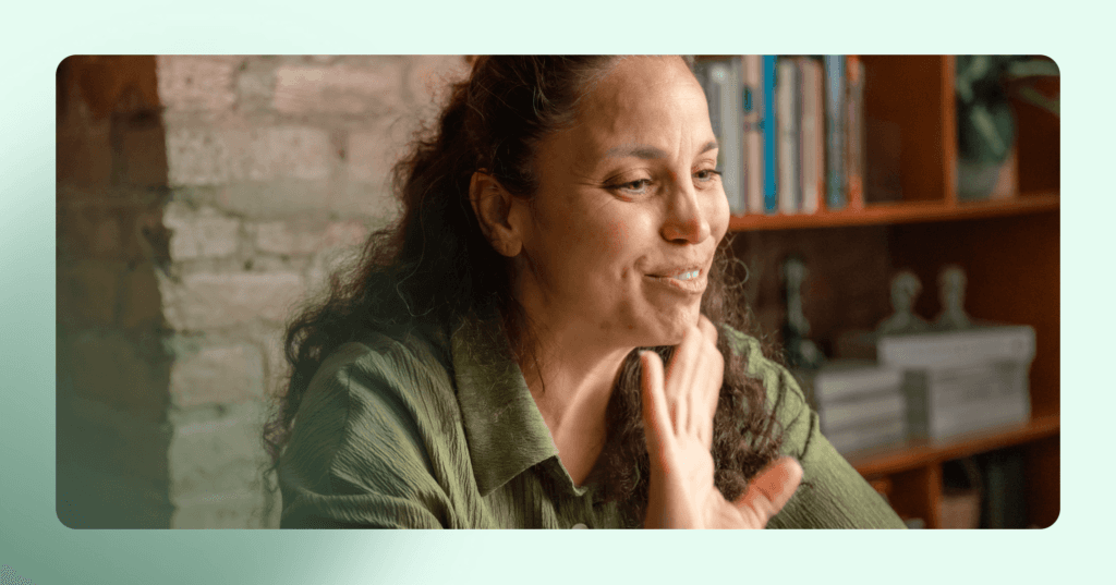 Woman in green shirt is sitting in front of a brick wall and waving. She has a smile on her face. The image is framed in light green.