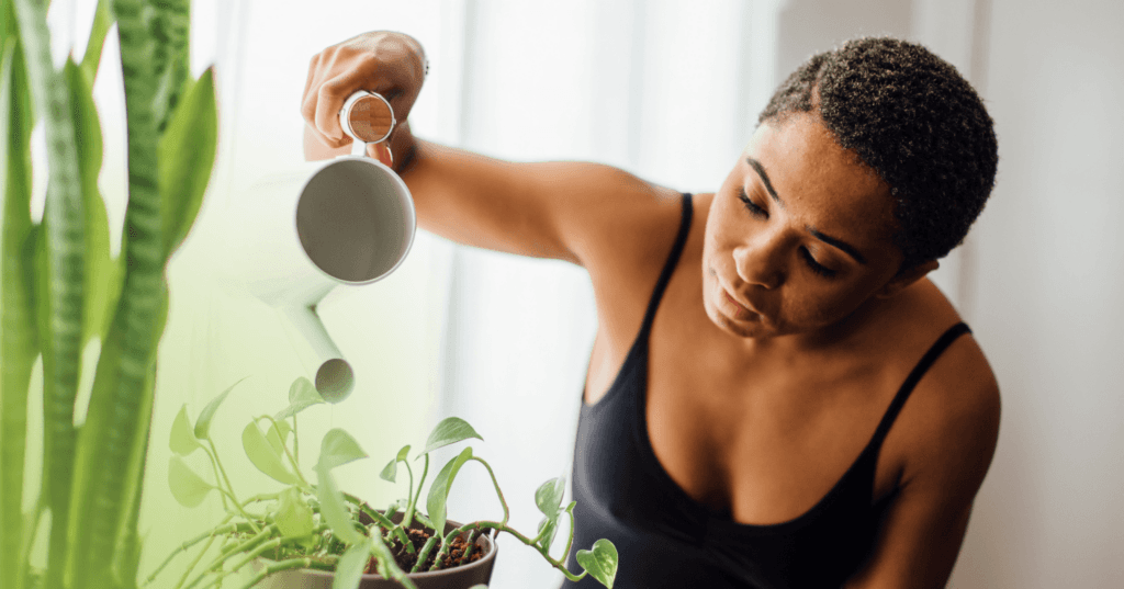 Woman is watering a plant with a white watering can. She is looking down at the plant. She is wearing a black tank top. The background is white.