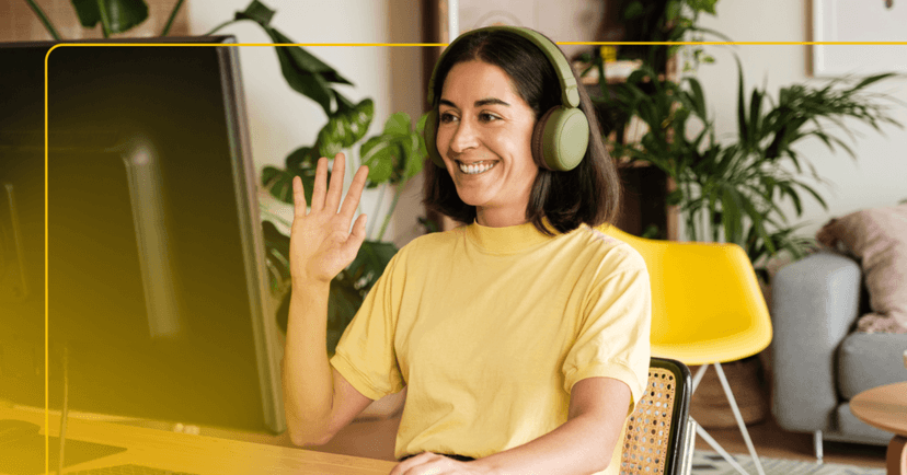 Woman is sitting at a desk in front of a desktop computer. She is looking at the computer screen smiling and waving her right hand. She is wearing a yellow T-shirt and green over the ear headphones. The background is her living room with a gray couch and multiple green plants.