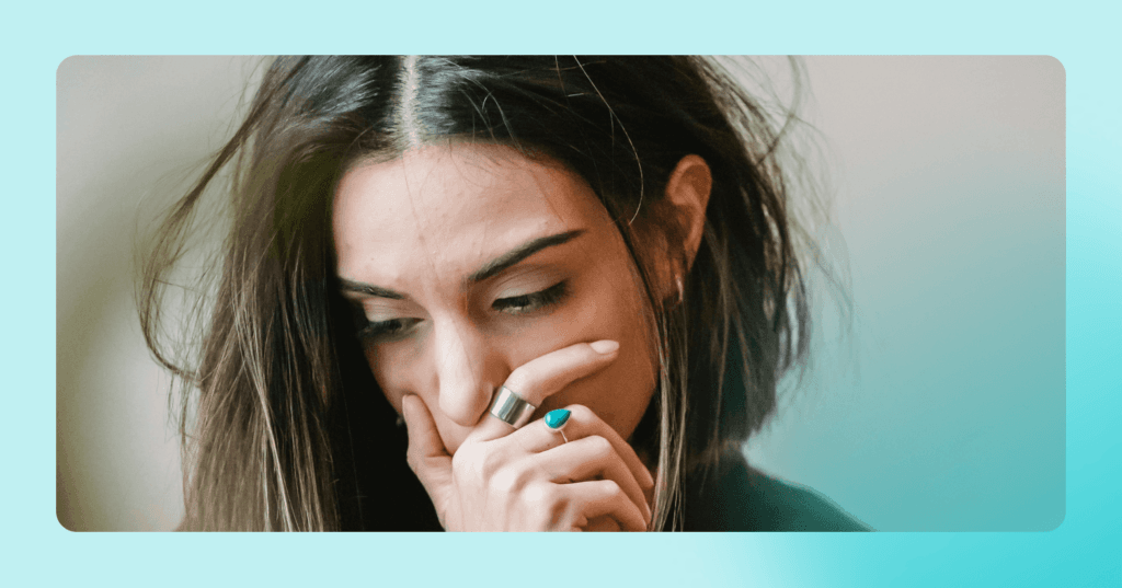 Woman stands looking down to her right. Her hand is covering her mouth. The wind is blowing her hair and she is wearing two rings. There is a blue border around the image.