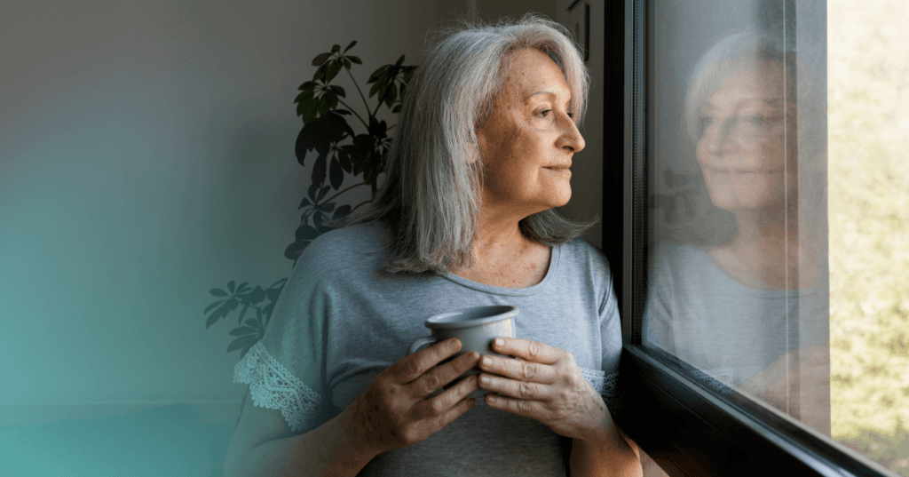 Woman stands indoors staring out the window. She is holding a mug with both hands in front of her. She is wearing a gray T-shirt and her reflection is visible against the window on the right. The background is a green plant against a white wall.